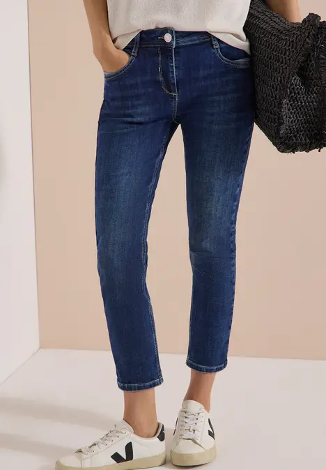 Person trägt dunkelblaue Jeans, weiße Sneakers und hält eine schwarze Tasche in der Hand.