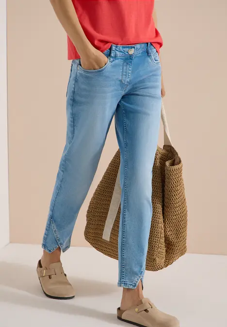 Frau trägt hellblaue Jeans mit Knöchelschlitz, rotes Shirt, Basttasche und helle Slipper.