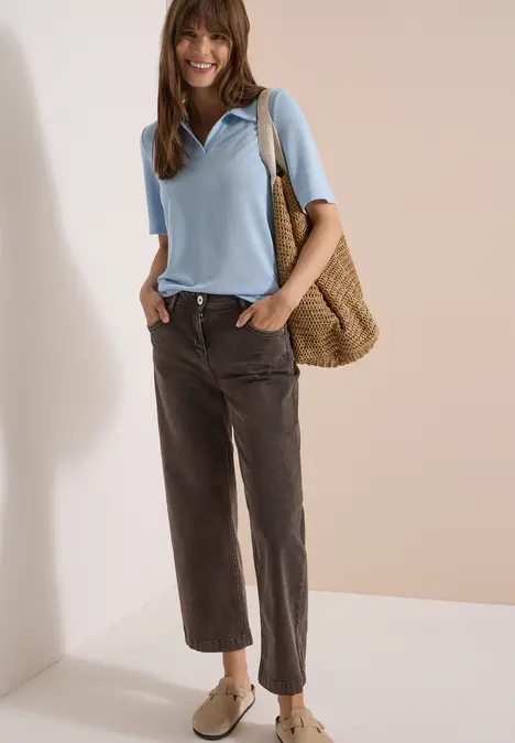 Frau in hellblauem Kurzarm-Pullover, braunen, weiten, verkürzten Hosen, mit Strohtasche über der Schulter und Loafers.