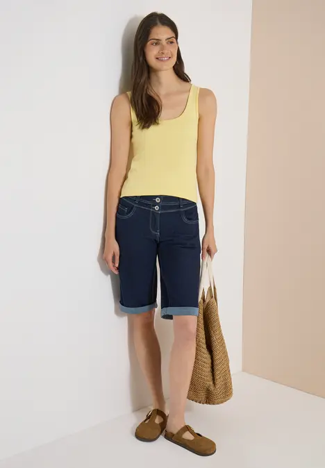 Frau in gelbem Top, Jeansbermuda, braunen Sandalen und Strohtasche vor hellem Hintergrund.