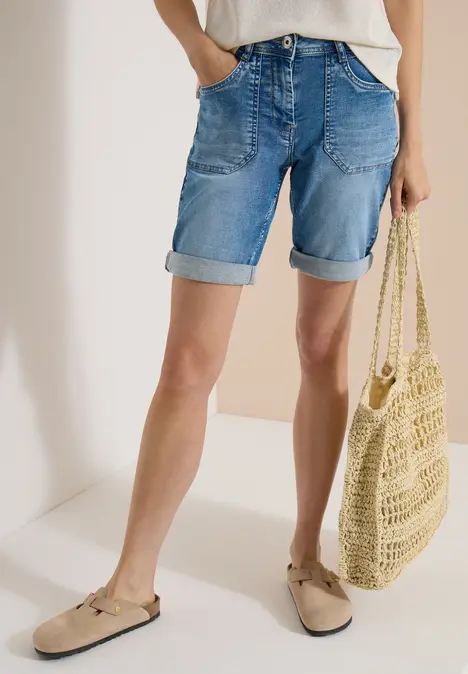 Frau in hellen Jeans-Shorts mit umgekrempelten Beinen, beigen Slippern und gehäkelter Strohtasche.