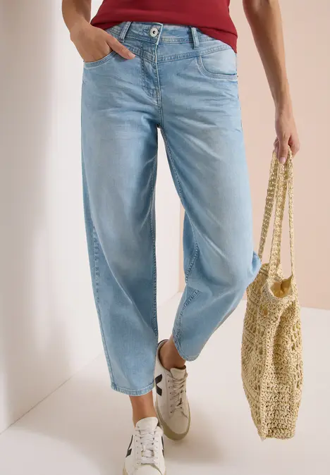 Person trägt hellblaue Jeans, weiße Sneaker und hält eine gewebte Tasche.