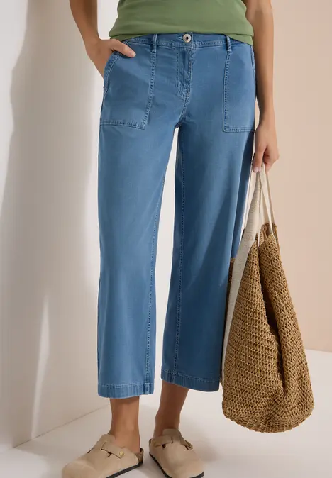 Frau trägt weite, knöchellange Jeans, grünes Oberteil, Strohtasche und beige Mules.