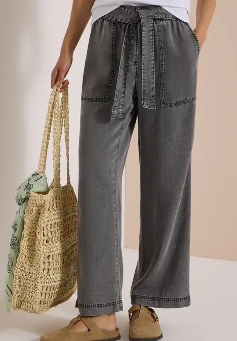 Graue, weite Bundfaltenhose mit Taillengürtel, aufgesetzten Taschen und Basttasche mit grünem Tuch.