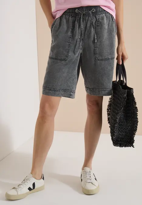 Person trägt ausgewaschene Jeans-Shorts mit Kordelzug, weiße Sneaker und schwarzen, geflochtenen Shopper.