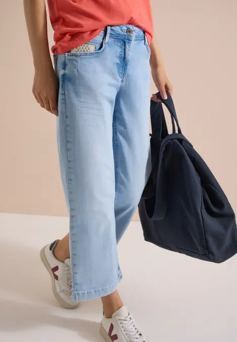 Person von der Taille abwärts in hellblauen, weiten Jeans mit weißen Sneakern und dunkelblauer Tasche.