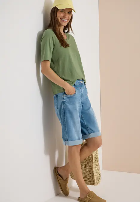 Junge Frau lehnt entspannt an der Wand, trägt grünes T-Shirt und knielange Jeans-Shorts.