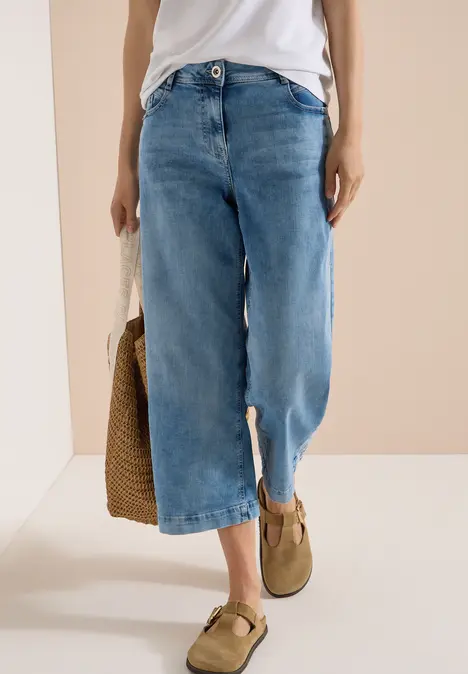 Frau in weißem Top trägt weite Culotte-Jeans mit Beinschlitz, beigen Loafern und hält eine geflochtene Tasche.