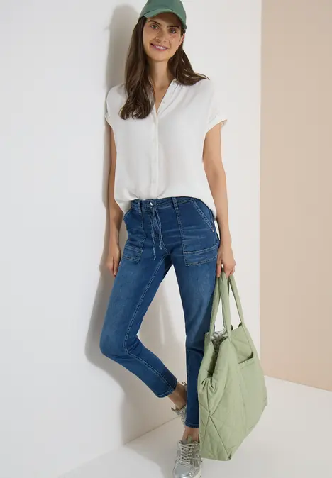 Frau in weißem T-Shirt, blauen Jeans und Baseballcap mit grünem Shopper, die entspannt an einer Wand lehnt.