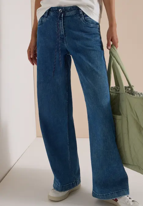 Locker sitzende blaue Jeans mit weitem Bein, weißem Top und grünem Stoffbeutel.