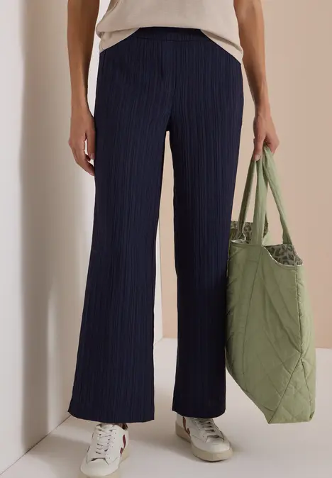 Frau in locker sitzender navy Nadelstreifenhose, weißem Shirt, weißen Sneakern und grüner, gesteppter Tasche.
