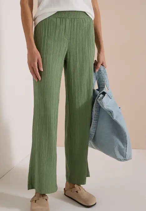 Ein Modell trägt grüne Plissee-Hose mit weitem Bein und hoher Taille, dazu weiße Schuhe und Jeansjacke.