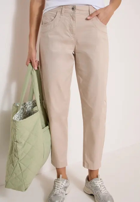 Eine Person trägt beige Chinohose, weiße Sneaker und hält eine grüne, gesteppte Tasche.