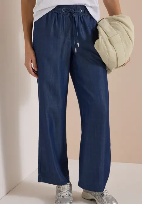 Dunkelblaue Jeanshose mit Kordelzug und geradem Bein, kombiniert mit Sneakern, dazu eine helle Steppjacke.