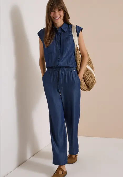 Frau in ärmellosem Jeans-Jumpsuit mit Strohtasche und Sandalen, die lächelnd posiert.
