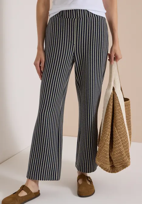 Frau trägt weiße Bluse, dunkelblaue Hose mit weißen Streifen, Strohtasche und braune Slipper.