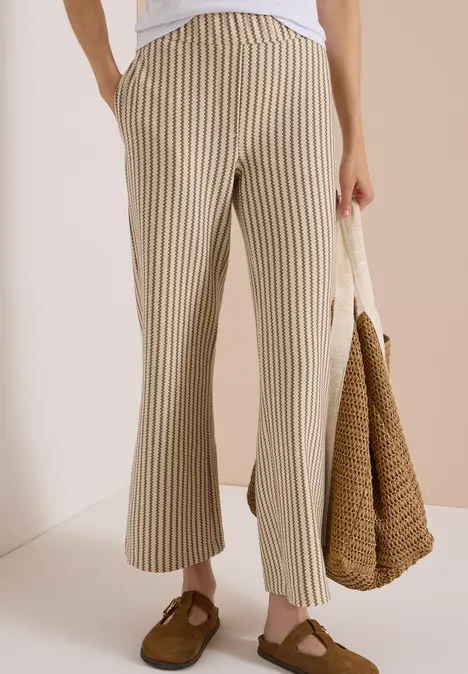 Beine in beigen, gestreiften Hosen, braune Loafers und eine Strohtasche in der Hand.