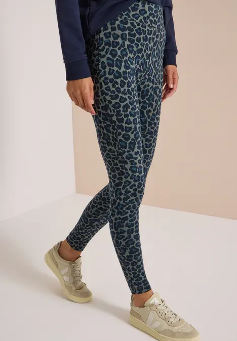 Beine einer Person in dunkelblauer Jacke, grün-blauen Leoparden-Leggings und beigen Sneakern vor hellem Hintergrund.