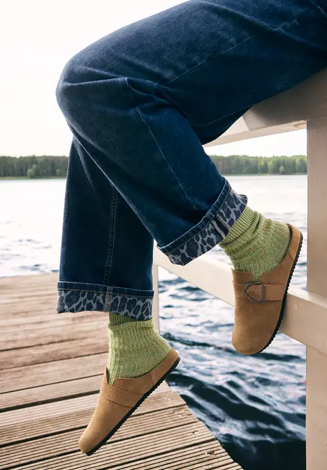 Person sitzt am Steg, trägt Jeans, grüne Socken und braune Hausschuhe am Seeufer.