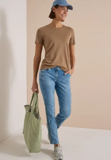 Frau in braunem T-Shirt, hellblauen Jeans, grauen Sneakers und blauer Kappe mit grünem Stoffbeutel vor beiger Wand.