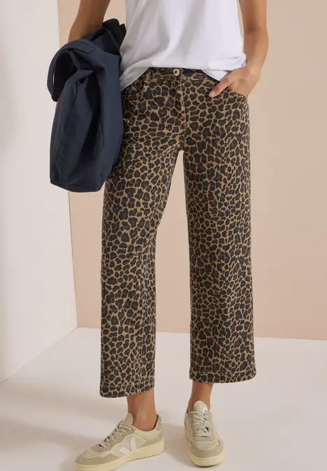 Eine Person trägt eine weiße Bluse, weite Leo-Hose und Sneaker vor beigem Hintergrund.