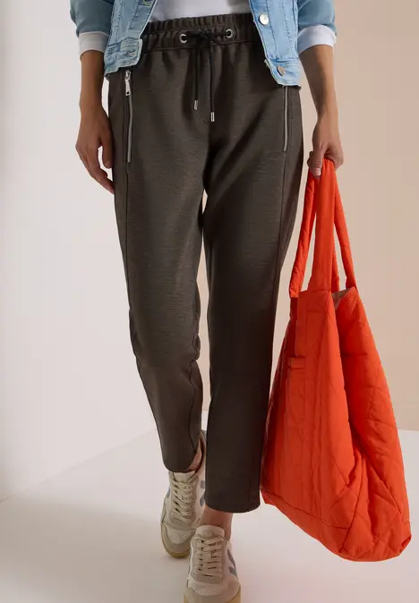 Person trägt Jeansjacke, weiße Bluse, schwarze Jogginghose, Turnschuhe und hält eine orangene Stepp-Tasche.