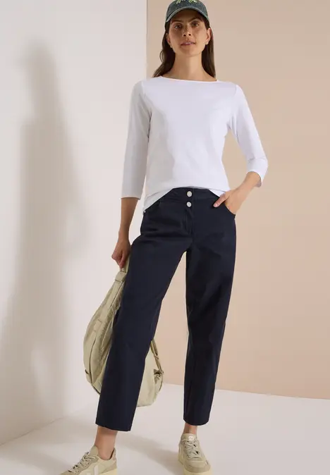Frau trägt weißes Shirt, dunkelblaue Hose, Sneaker und Cap, hält Jacke lässig über die Schulter.