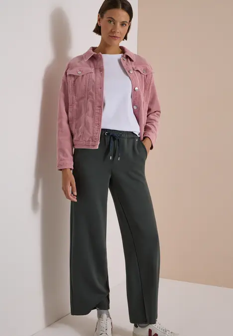 Frau vor neutralem Hintergrund mit rosa Jeansjacke, weißem Shirt, weiten schwarzen Hosen und weißen Sneakern.