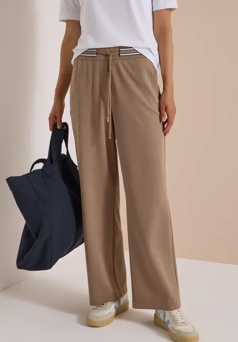 Person trägt weißes T-Shirt, weite beige Hose, weiße Sneaker und hält große dunkelblaue Tasche vor neutralem Hintergrund.