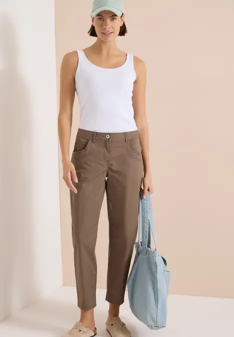 Frau in weißem Tanktop und brauner Hose mit blauer Tasche vor hellem Hintergrund.