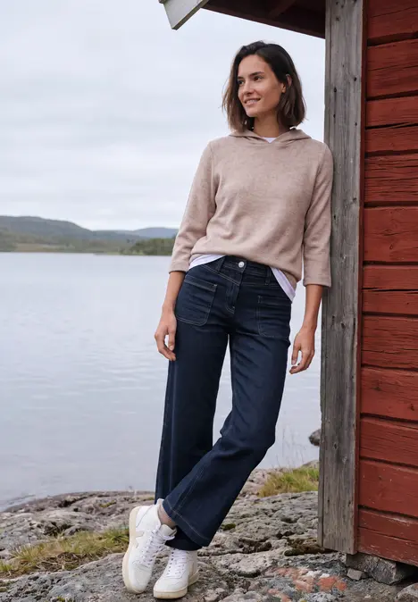 Frau lehnt lächelnd an roter Holzhütte am See, trägt Pullover, Jeans und weiße Sneakers, im Hintergrund Natur.
