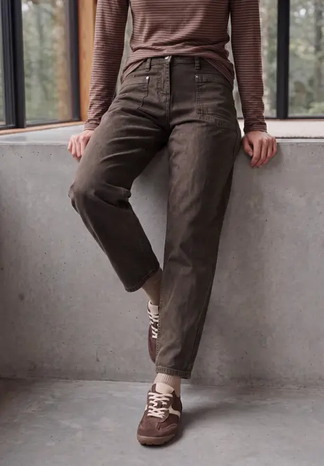 Dunkelbraune, hoch taillierte Cordhose und passende Sneaker, Person lehnt an einer Betonbrüstung.