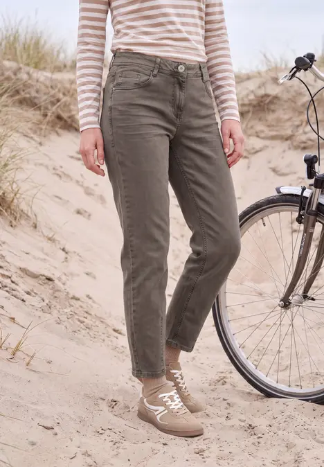 Person in olivgrünen Hosen, gestreiftem Langarmshirt und Sneakern steht im Sand neben einem Fahrrad.