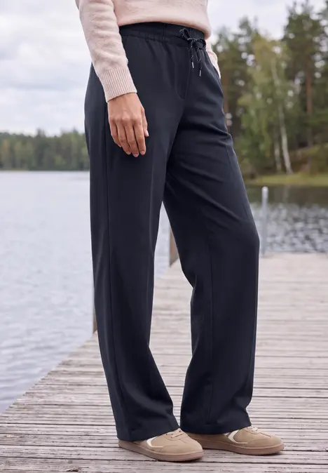 Frau in bequemen, weiten schwarzen Hosen und Hausschuhen steht auf einem Pier vor einem Waldsee.
