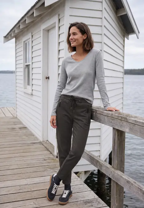 Frau im grauen Shirt und dunkler Hose lehnt lächelnd an einem Steg mit weißer Hütte und Wasser im Hintergrund.