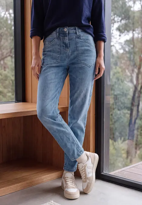 Benen van een persoon in blauwe jeans en witte sneakers, staand voor een knus raam met boszicht.