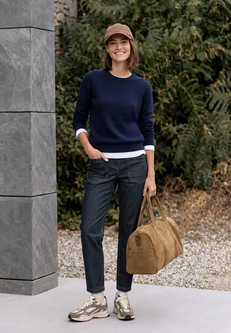 Eine Frau steht draußen in marineblauem Pullover, Jeans und Sneakern, hält eine braune Reisetasche.