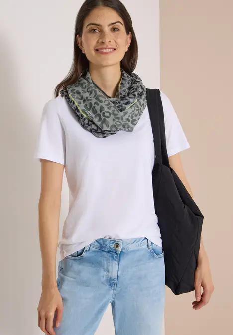 Eine lächelnde Frau trägt weißen T-Shirt, blaue Jeans, grünen Schal und schwarze Umhängetasche.