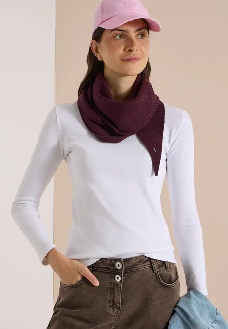 Frau mit weißem Langarmshirt, beiger Hose, violettem Dreiecksschal und rosa Cap, hält einen blauen Pullover