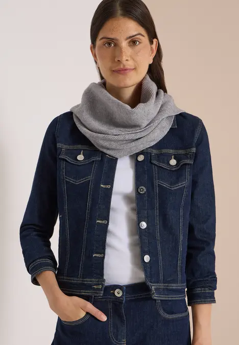 Frau mit dunklen Haaren trägt blaue Jeansjacke und grauen Schal.