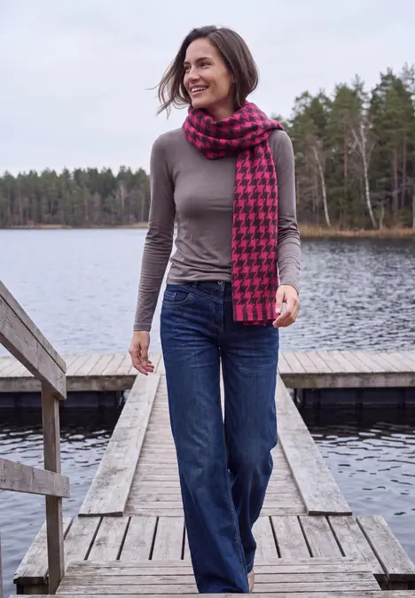 Frau spaziert lächelnd auf einem Steg am See, trägt graues Oberteil, Jeans und rot-schwarzen Hahnentritt-Schal vor Waldkulisse.