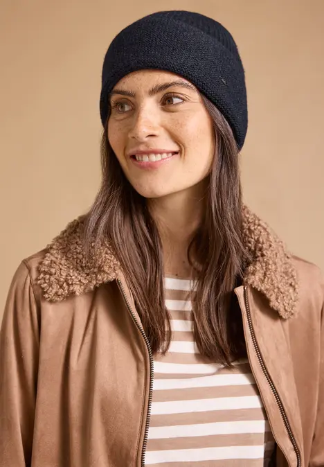 Frau mit schwarzer Strickmütze, brauner Jacke mit Teddy-Kragen und gestreiftem Shirt vor beigem Hintergrund.