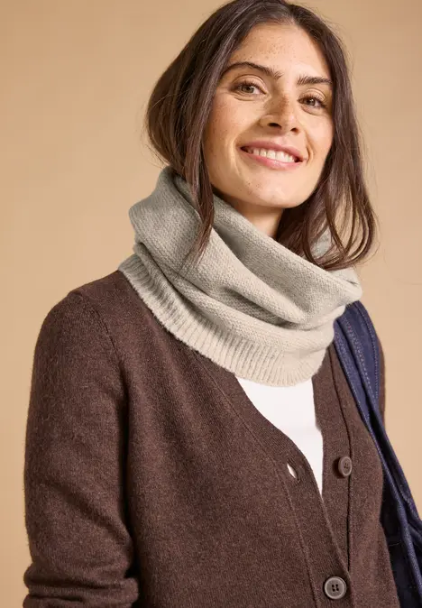Lächelnde Frau mit brauner Strickjacke und beigem Schlauchschal vor hellem Hintergrund.