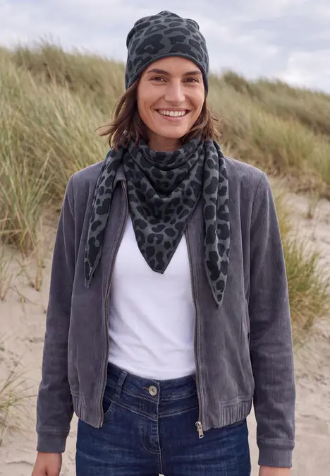 Person steht auf einem Dünenstrand, trägt weißes T-Shirt, graue Jacke, gemusterten Schal und Mütze, lächelt.
