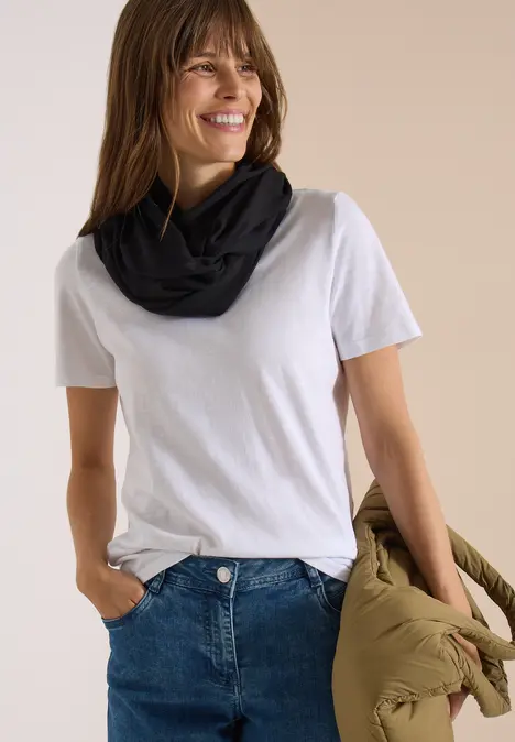 Frau trägt weiße T-Shirt, Jeans und schwarzen Loop-Schal, hält eine olivgrüne Jacke und lächelt.