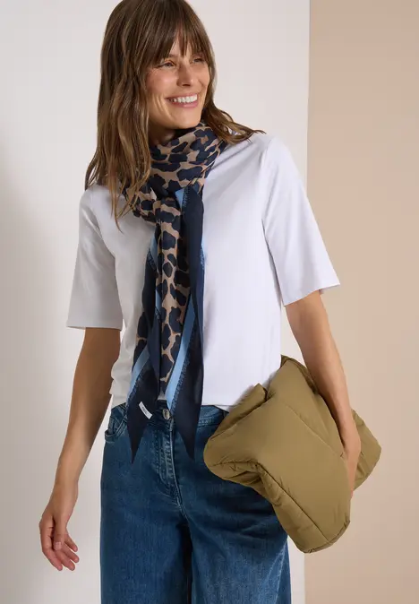 Frau in weißem T-Shirt mit blau gemustertem Schal und grünem Kissen in der Hand.