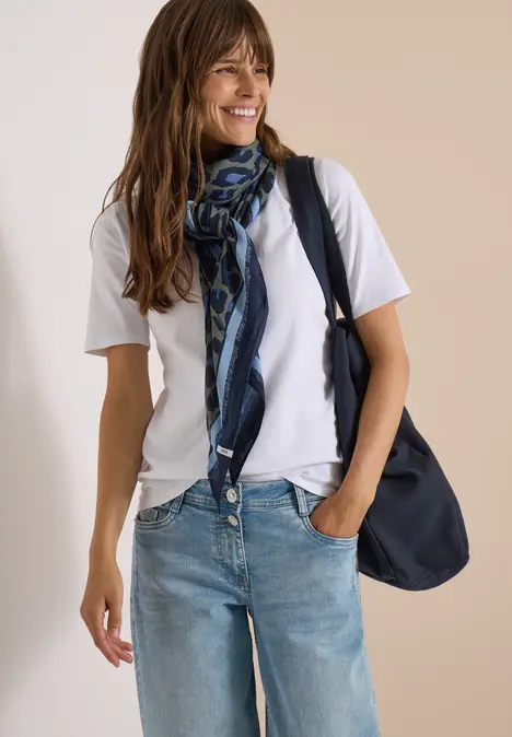 Frau in weißem T-Shirt, Jeans und blauem Schal trägt lässige Tasche und lächelt.