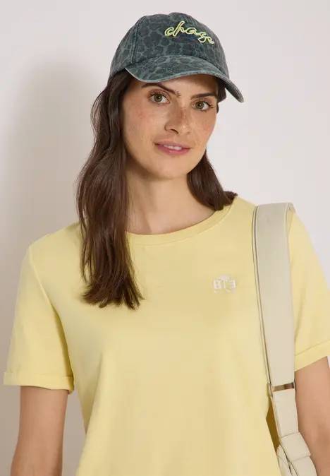 Frau mit Jeans-Cap, gelbem T-Shirt und Schultertasche, die in die Kamera lächelt.