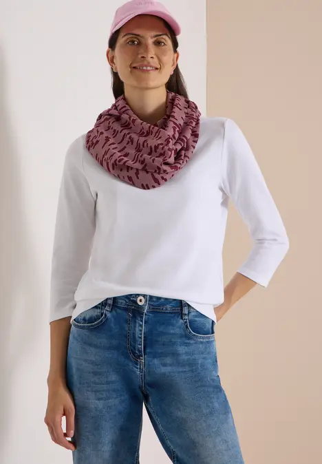 Frau mit rosa Cap, gemustertem Schlauchschal, weißem Shirt und blauen Jeans steht lächelnd.