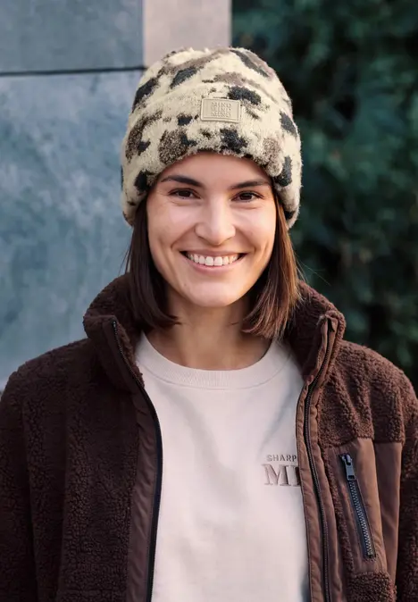 Junge Frau trägt einen camouflage-Bommelmütze und braune Fleecejacke, lächelt in die Kamera.